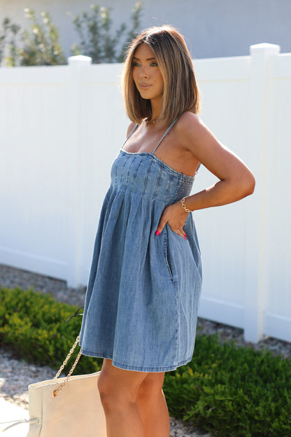 A woman in a Denim Pleated Babydoll Mini Dress stands outdoors by a white fence, hand in pocket, with a beige bag at her side.