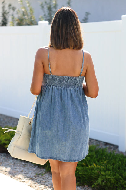 A woman in a Denim Pleated Babydoll Mini Dress carries a cream bag while walking outdoors near a white fence.