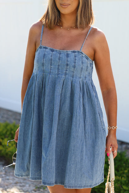 Woman wearing a sleeveless Denim Pleated Babydoll Mini Dress with spaghetti straps, holding sunglasses and a purse outdoors.