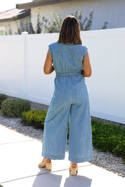 A woman stands outdoors, facing away, in a Denim Button Up Wide Leg Jumpsuit and beige sandals.