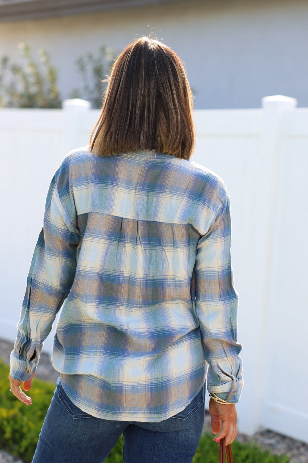 A person with shoulder-length hair in a Blue Plaid Button Down Flannel Shirt and jeans stands outside facing a white fence.