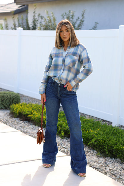 A woman stands outdoors in Medium Wash Flare Denim Jeans, holding a brown purse by some greenery.