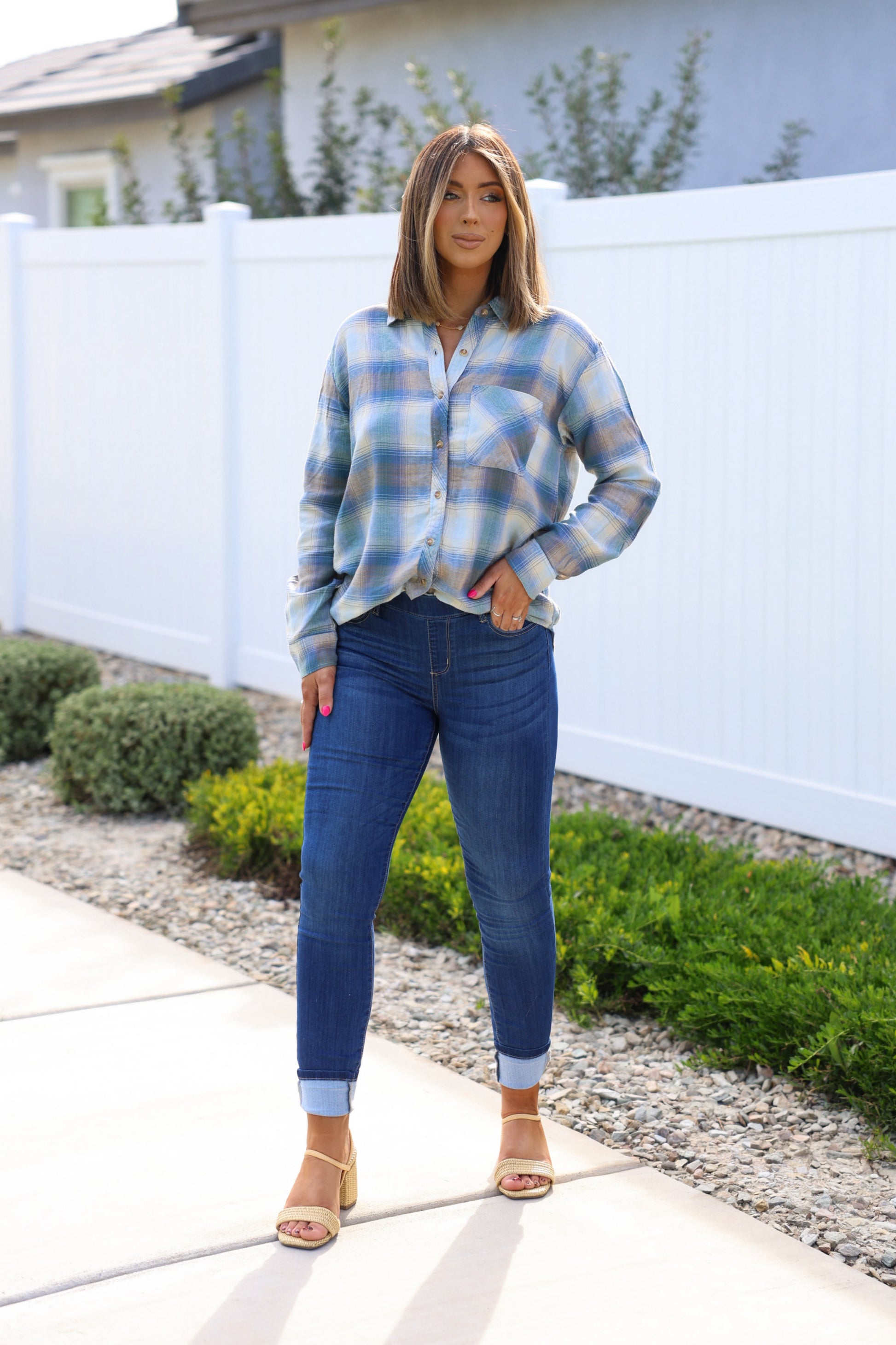 Woman outdoors by a white fence, in a plaid shirt, beige heeled sandals, and Dark Wash Pull On Cuffed Skinny Jeans.