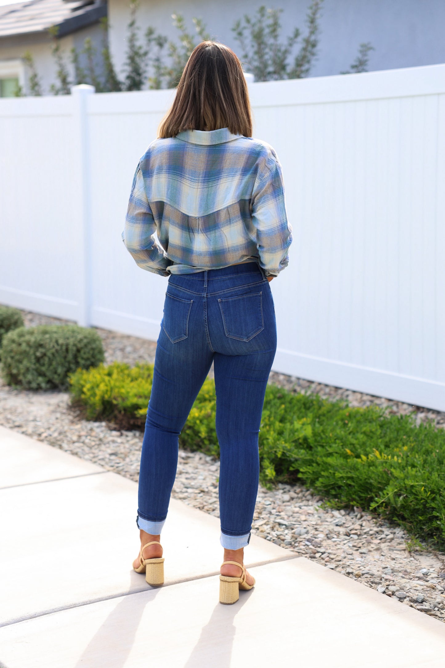 A woman stands outdoors with her back to the camera, wearing a plaid shirt and Dark Wash Pull On Cuffed Skinny Jeans.