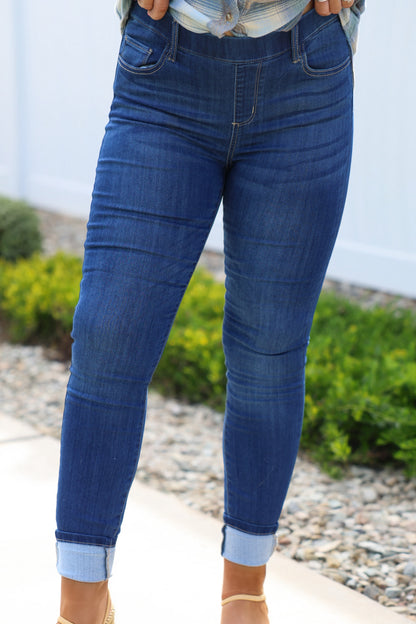 Person wearing Dark Wash Pull On Cuffed Skinny Jeans and a plaid shirt, standing on a sidewalk with greenery and rocks behind.