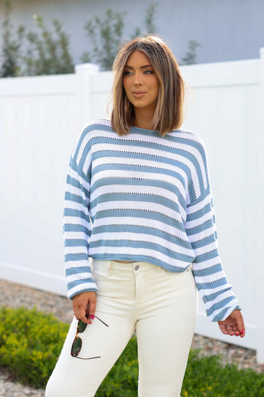 A woman outdoors in a Cloud Grey and White Striped Sweater and white pants, holding sunglasses by a white fence.