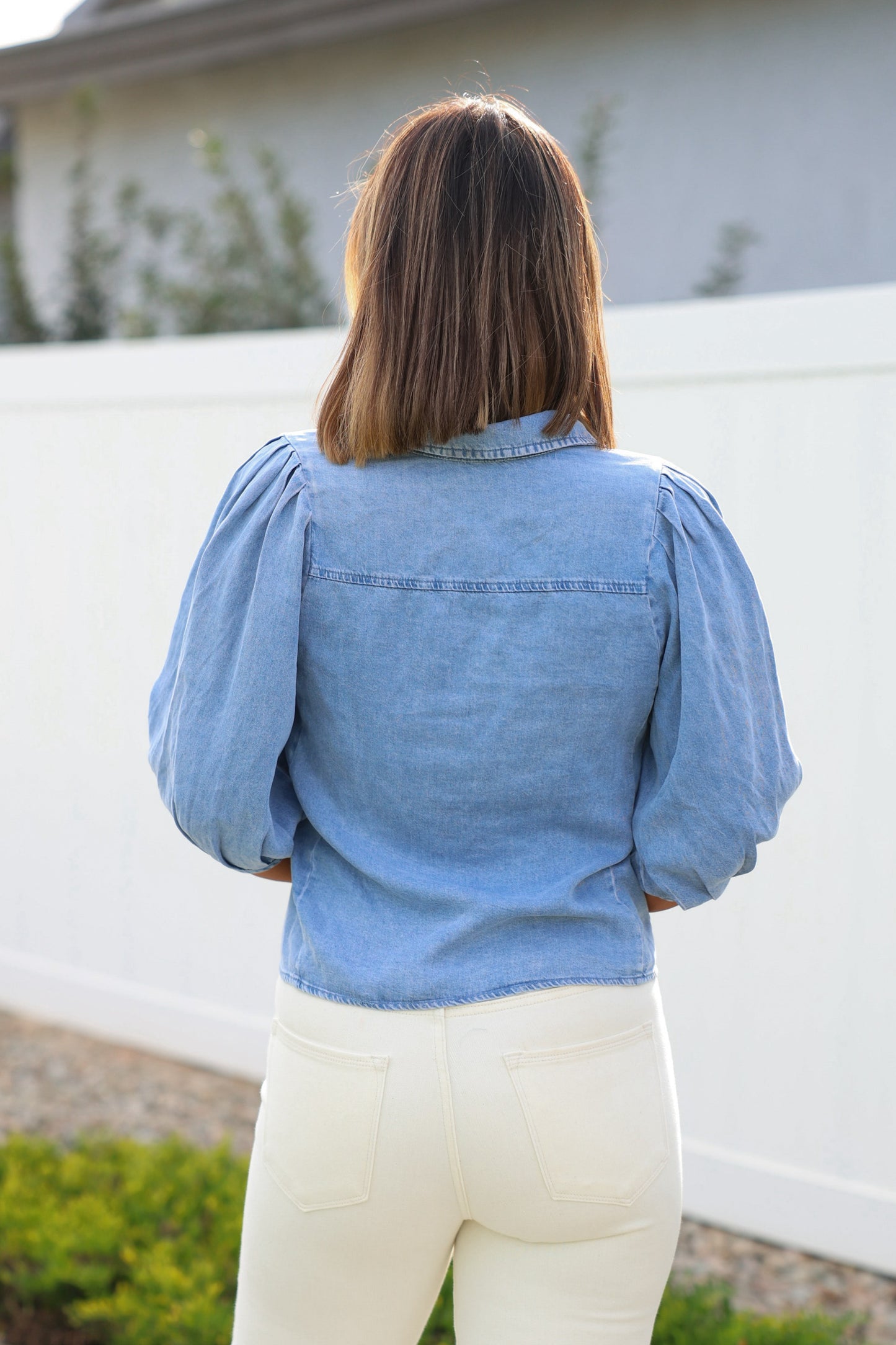 A woman with shoulder-length hair stands outside facing away, wearing a Light Denim Pleated Button Down Shirt.