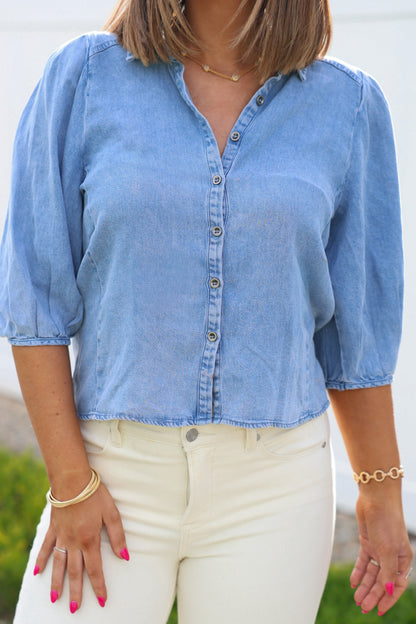 Woman wearing a Light Denim Pleated Button Down Shirt with ivory pants, gold bracelets, and pink nails outdoors.