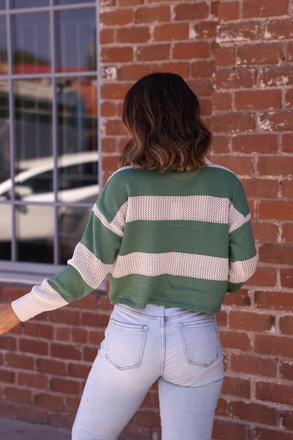 A person in a Declan Beige & Green Striped Sweater and light blue jeans stands facing a brick wall, shown from the back.
