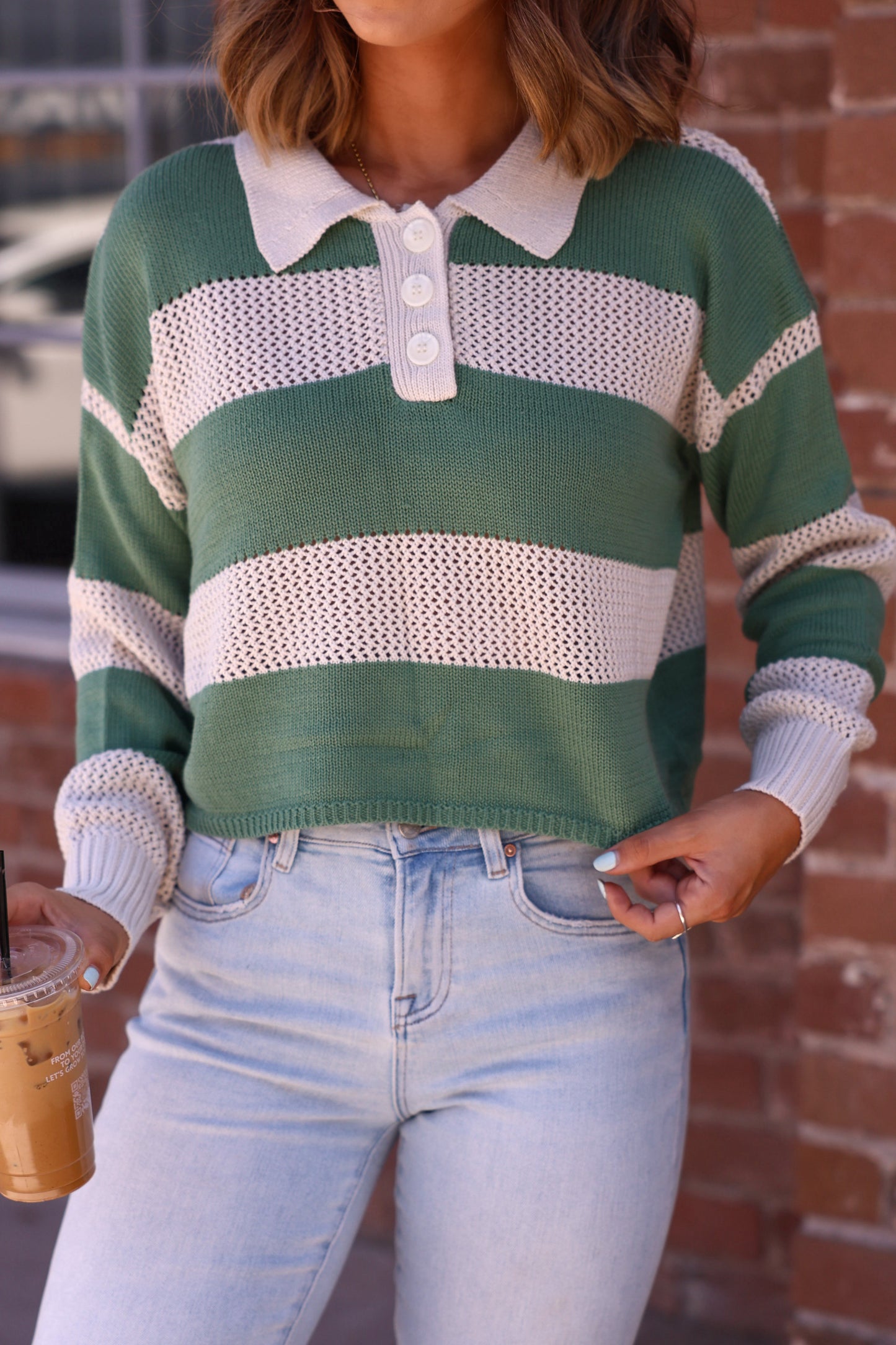 Woman in the Declan Beige & Green Striped Sweater with light jeans, holding iced coffee by a brick wall.