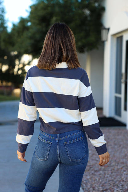 A woman stands outdoors with her back to the camera, wearing a Thread and Supply Navy Stripe Button Up Top and jeans.