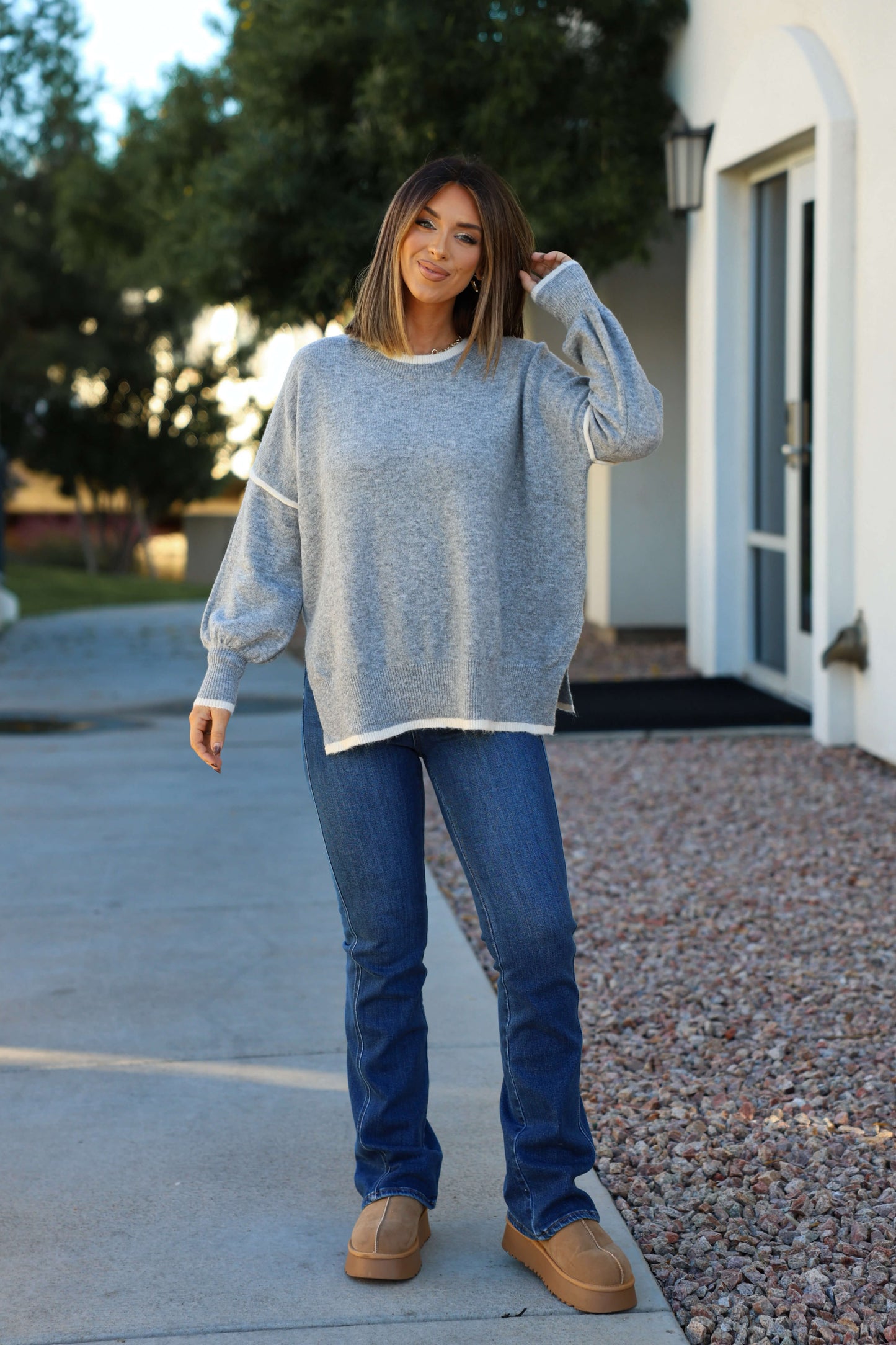 Woman standing outside in a Grey Contrast Detail Pullover Sweater, blue jeans, and tan shoes, smiling with one hand touching her hair.