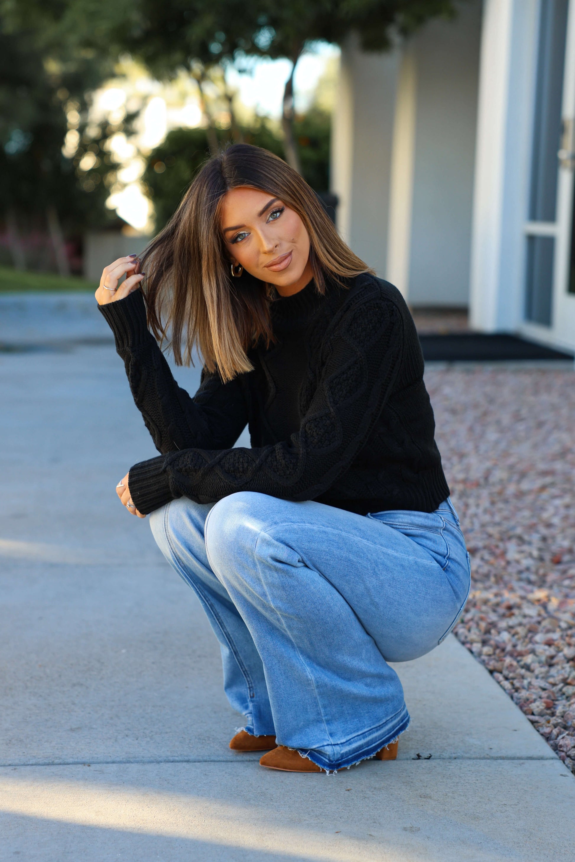 A woman with straight brown hair squats on a sidewalk, wearing a Chunky Black Mock Neck Sweater, light blue jeans, and brown shoes.