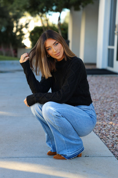 A woman with straight brown hair squats on a sidewalk, wearing a Chunky Black Mock Neck Sweater, light blue jeans, and brown shoes.