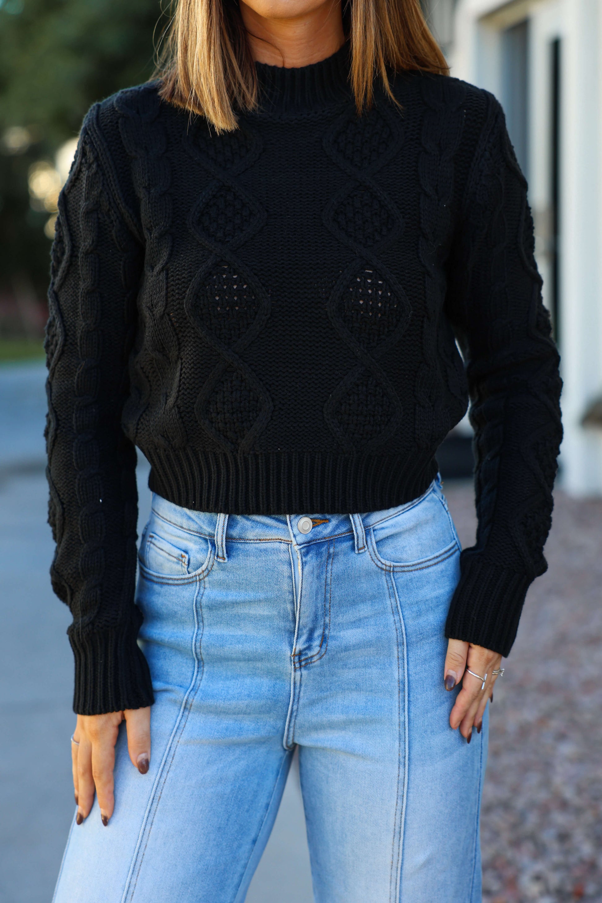 Woman in a chunky black mock neck sweater and high-waisted light blue jeans, standing outdoors with hands at her sides.