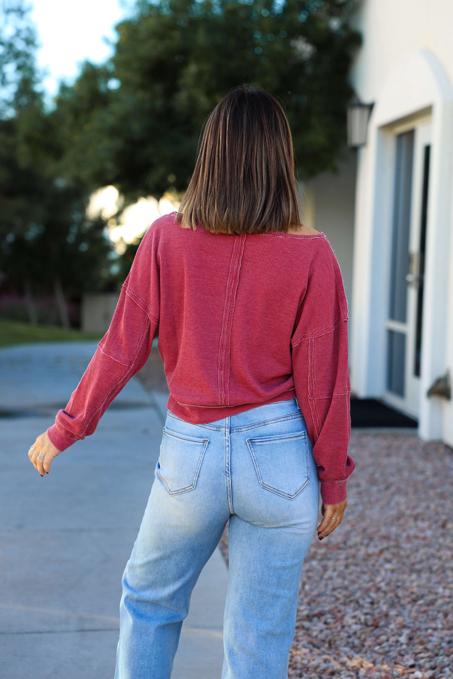 A woman with straight brown hair wears a Red Waffle Knit Boatneck Pullover and light blue jeans, standing outside facing away.