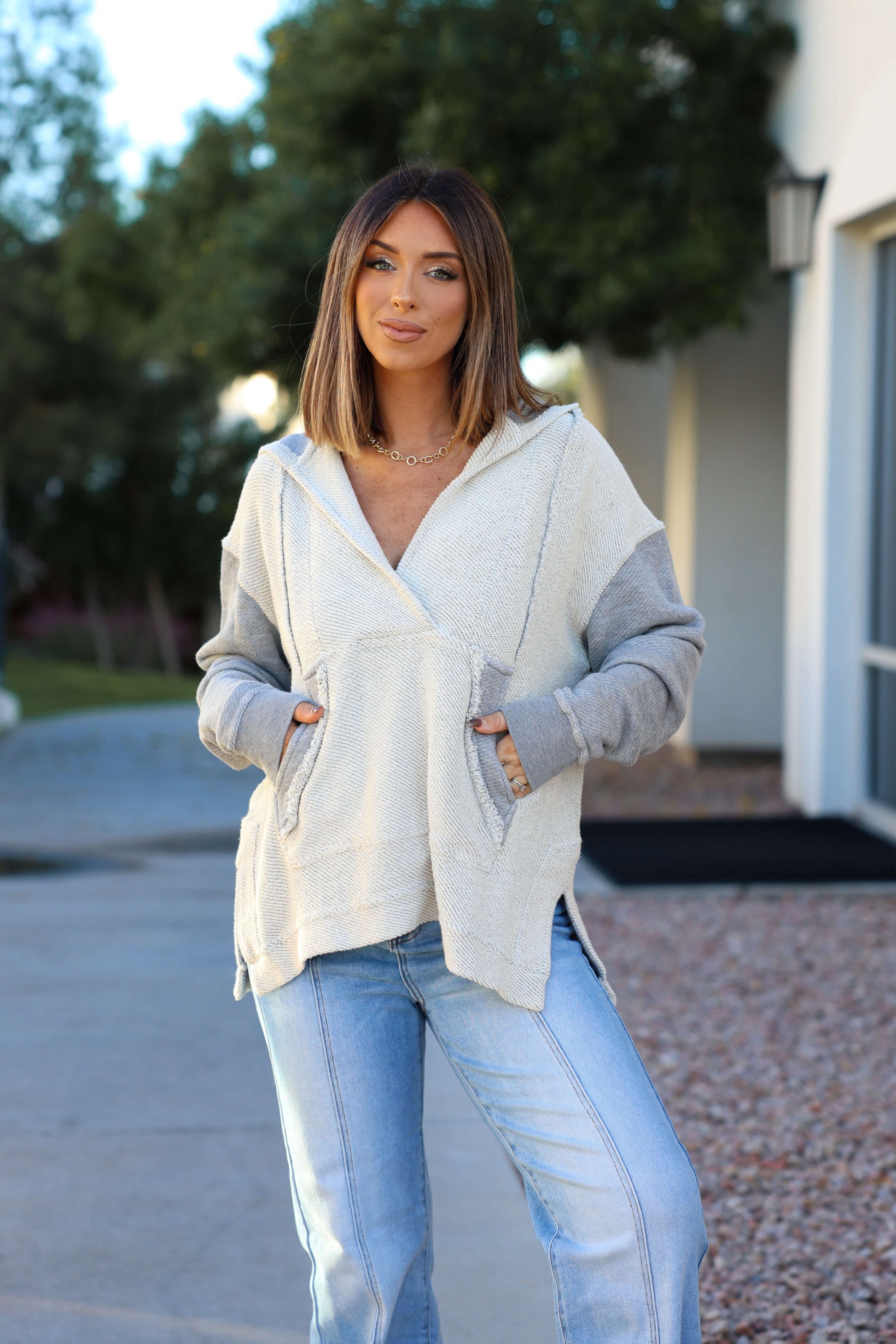 Woman with straight brown hair wears a Heather Grey Slouchy Hooded Sweatshirt and jeans, smiling outdoors with hands in her pockets.