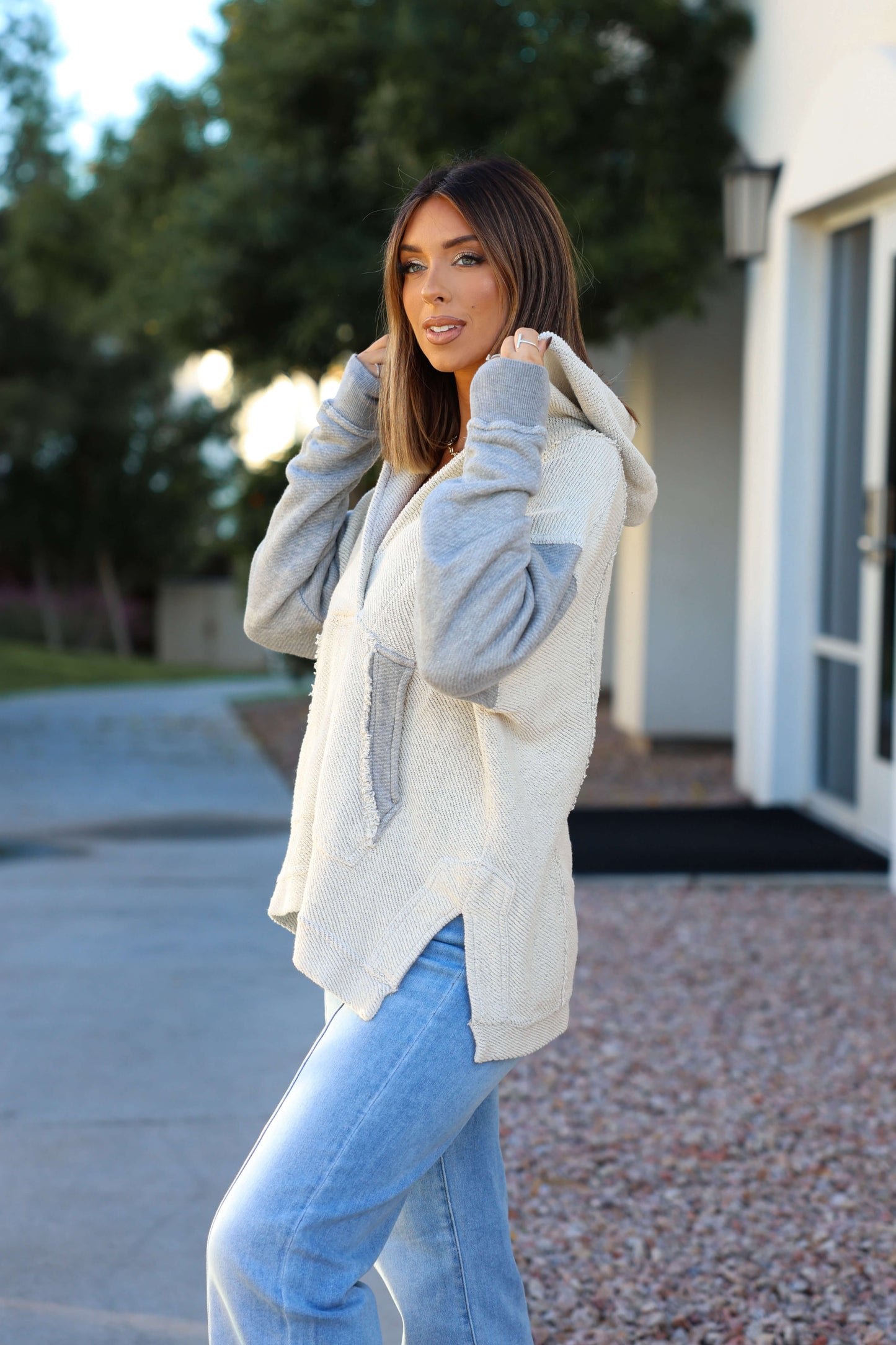 A woman in a Heather Grey Slouchy Hooded Sweatshirt and light blue jeans stands by a modern building, looking at the camera.