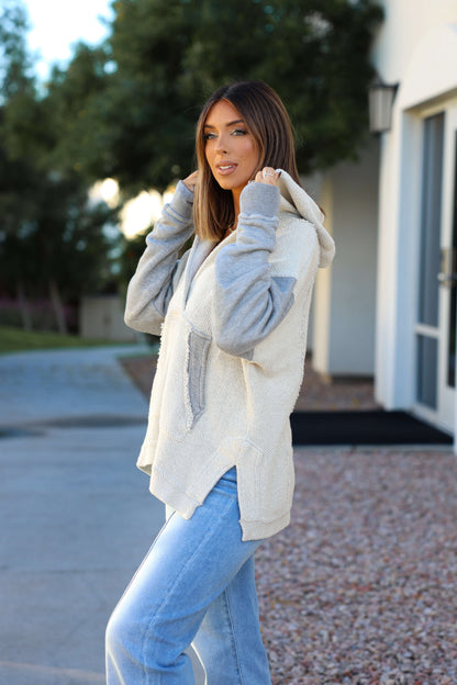 A woman in a Heather Grey Slouchy Hooded Sweatshirt and light blue jeans stands by a modern building, looking at the camera.