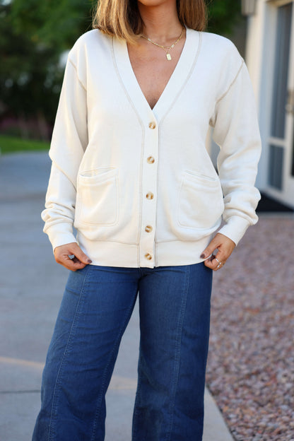 Woman wears the Thread and Supply Cream Stitch Buttoned Cardigan with gold buttons and blue jeans, standing outdoors.