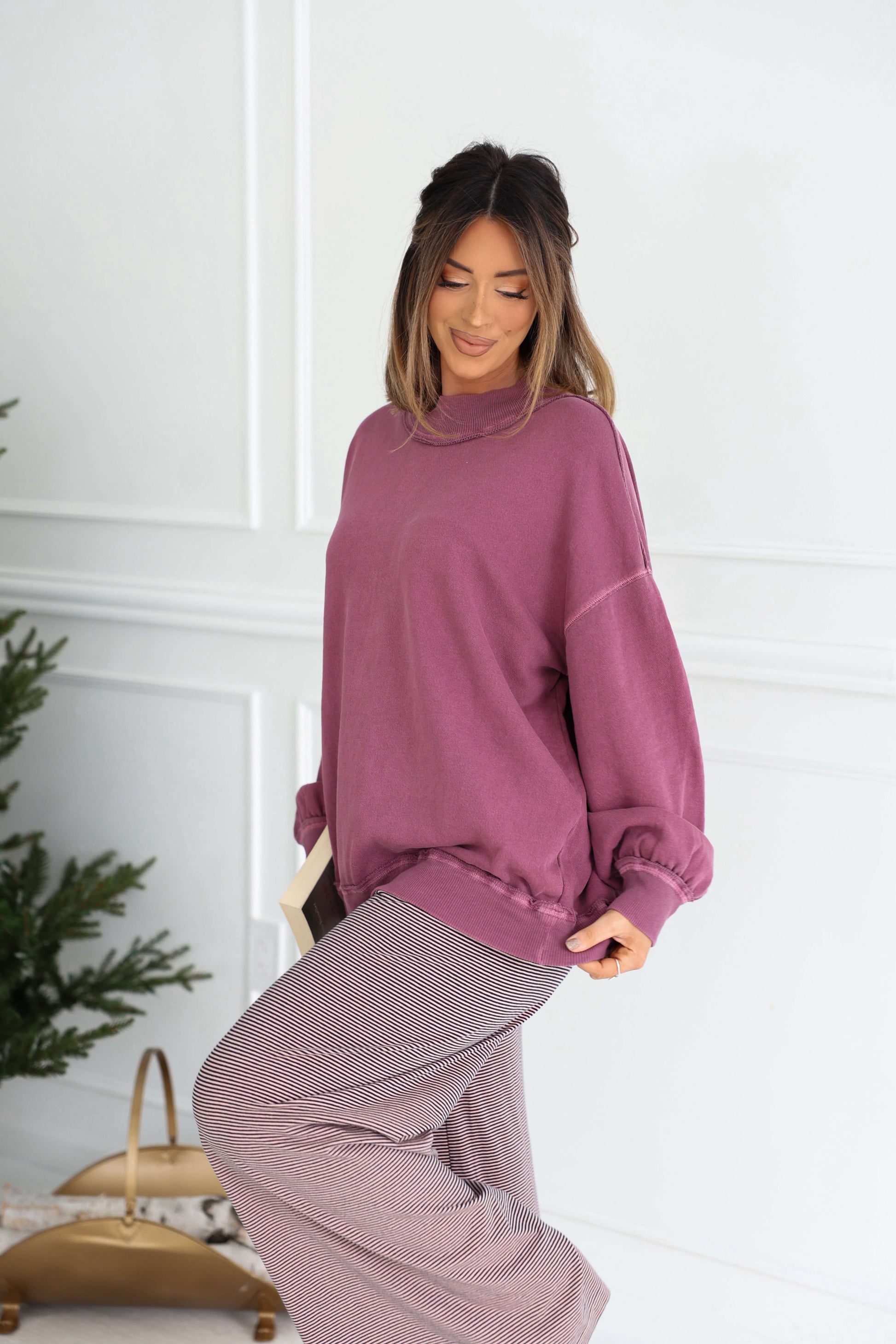A woman in a Free People Iris Orchid Palm Springs Pullover stands indoors holding a book, with a Christmas tree behind her.