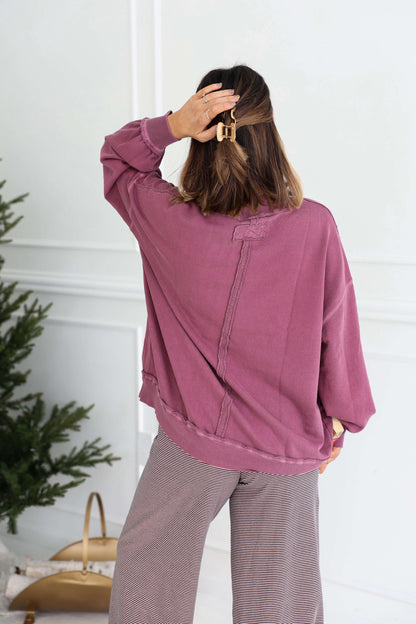 A woman in a Free People Iris Orchid Palm Springs Pullover and gray pants stands with her back to the camera, hand in her hair.