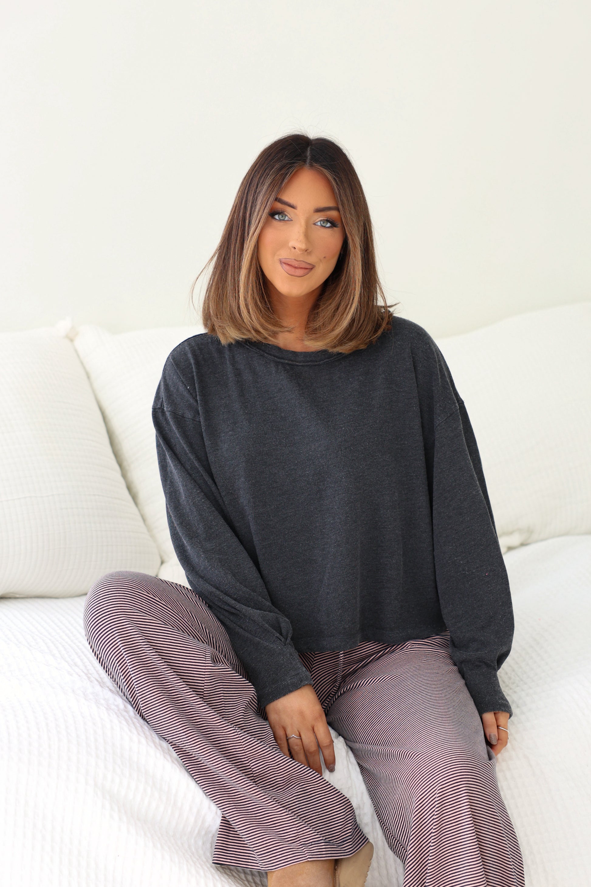 Woman with shoulder-length hair in a Charcoal Mineral Wash Long Sleeve Top and striped pants, sitting on a white bed.
