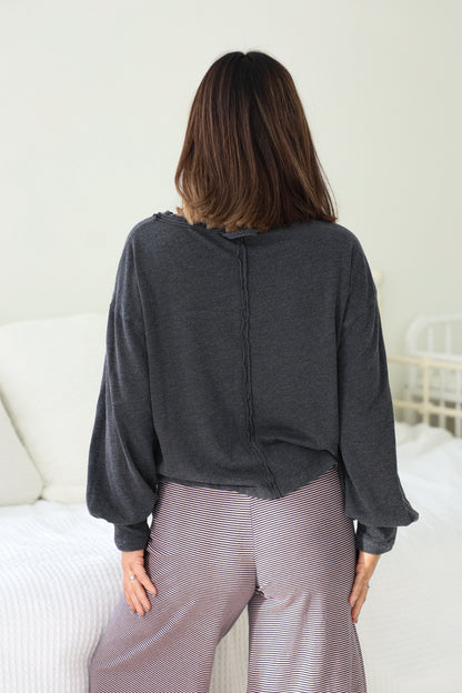 A woman with shoulder-length brown hair models the Charcoal Mineral Wash Long Sleeve Top, standing in a bedroom with her back to the camera.