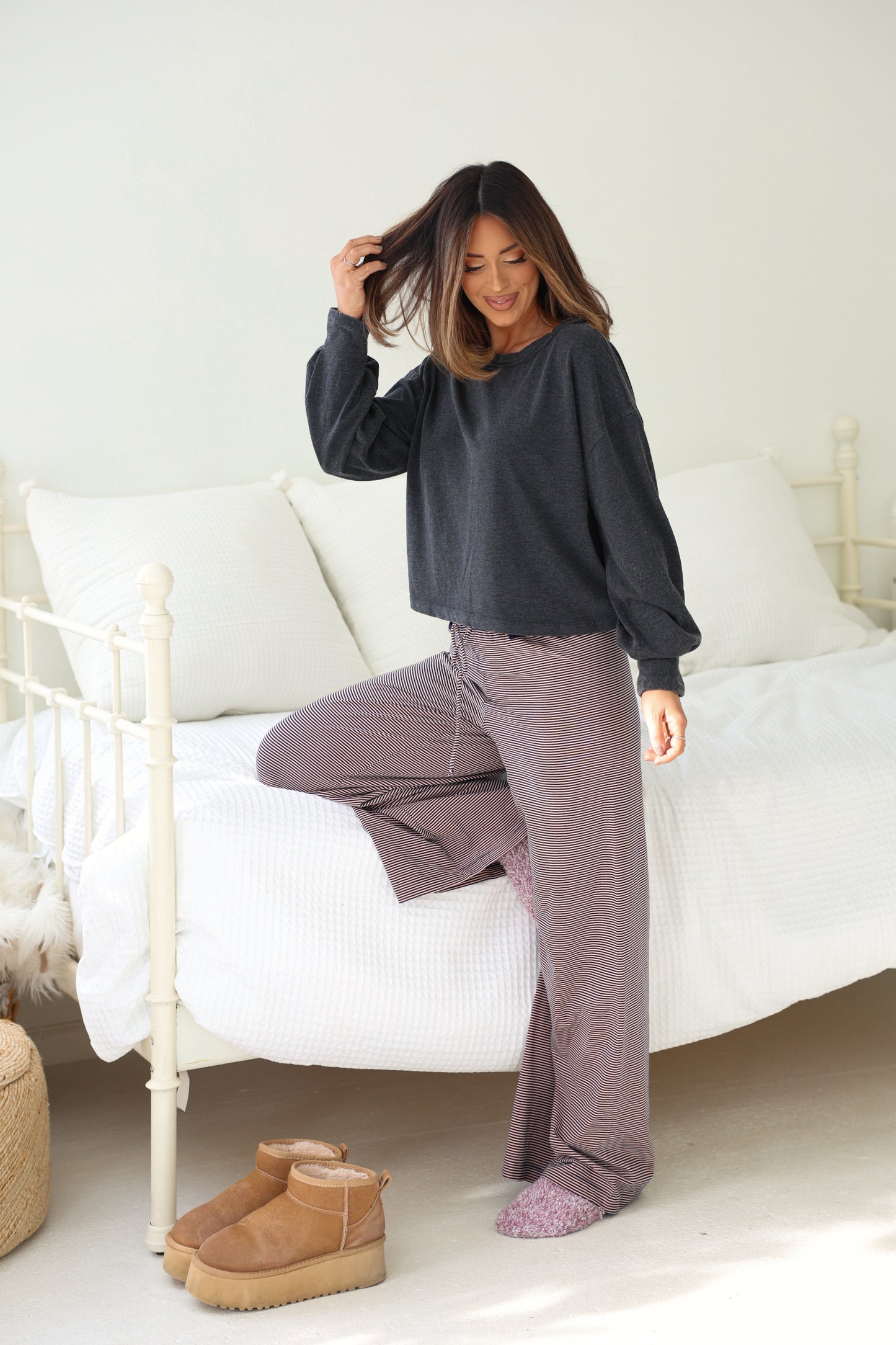 Woman smiles by a white bed, touching her hair in a Charcoal Mineral Wash Long Sleeve Top; slippers rest nearby.