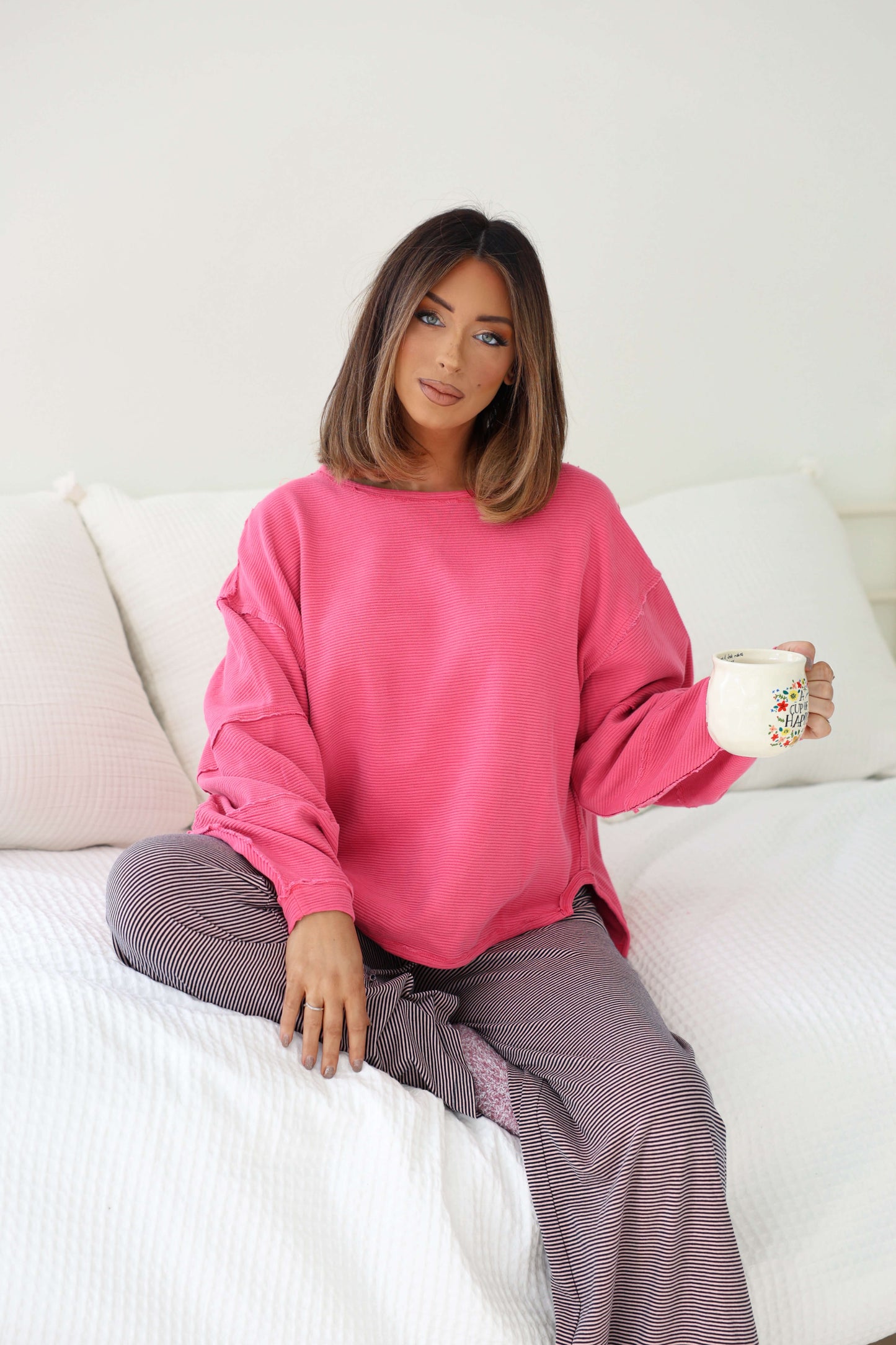 Woman with shoulder-length brown hair sits on a bed, wearing the Free People Pink Total Eclipse Top and striped pants, holding a mug.
