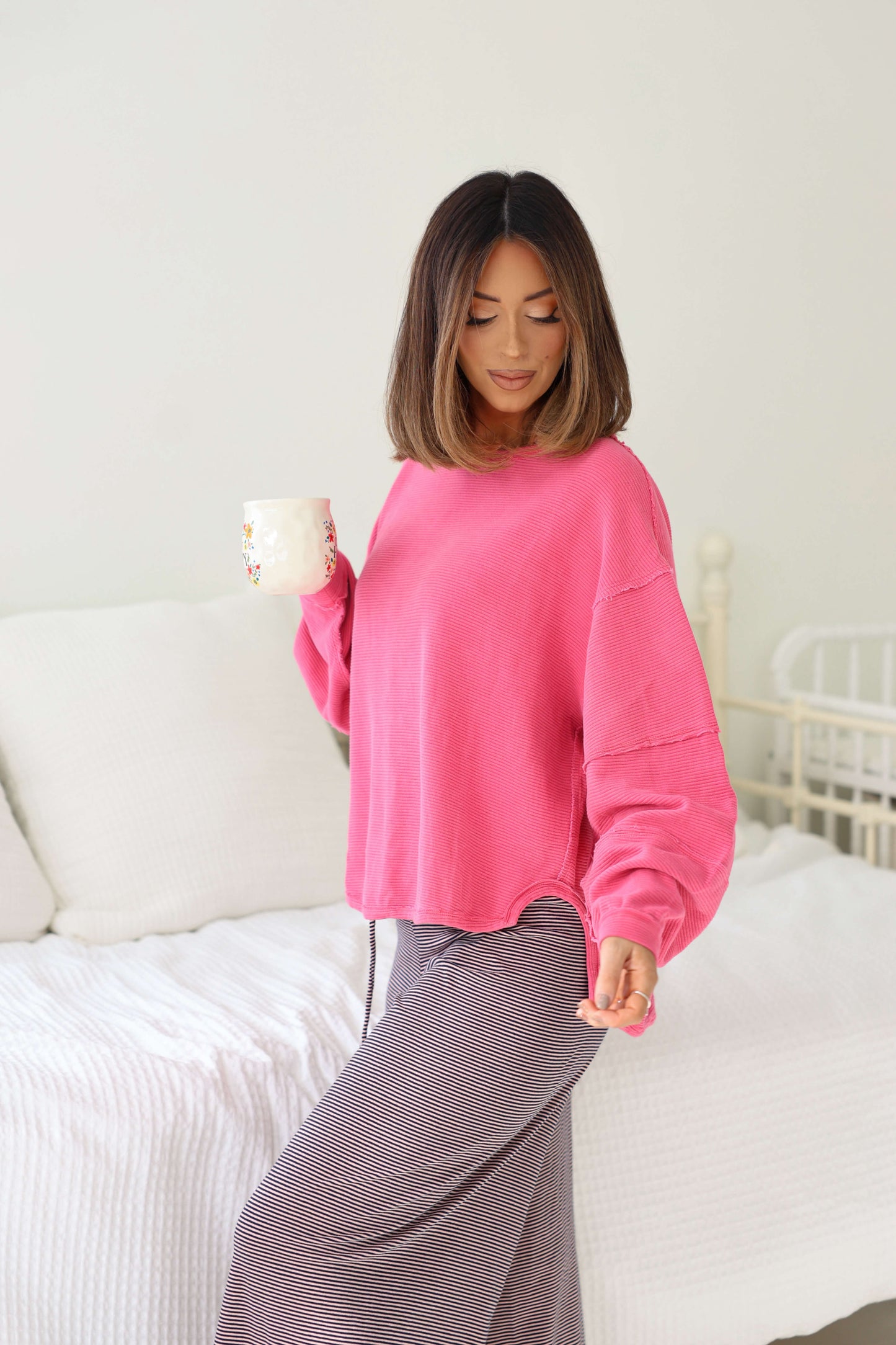 A woman in a Free People Pink Total Eclipse Top holds a mug and stands by a bed with white bedding, looking down and smiling.