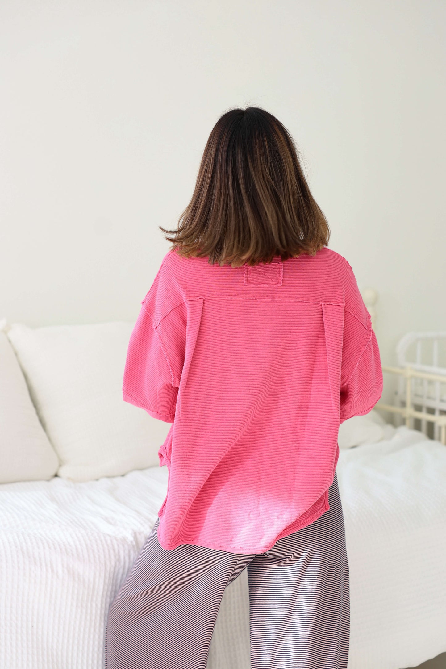 A person in a Free People Pink Total Eclipse Top and gray pants stands facing away from the camera near a white bed.