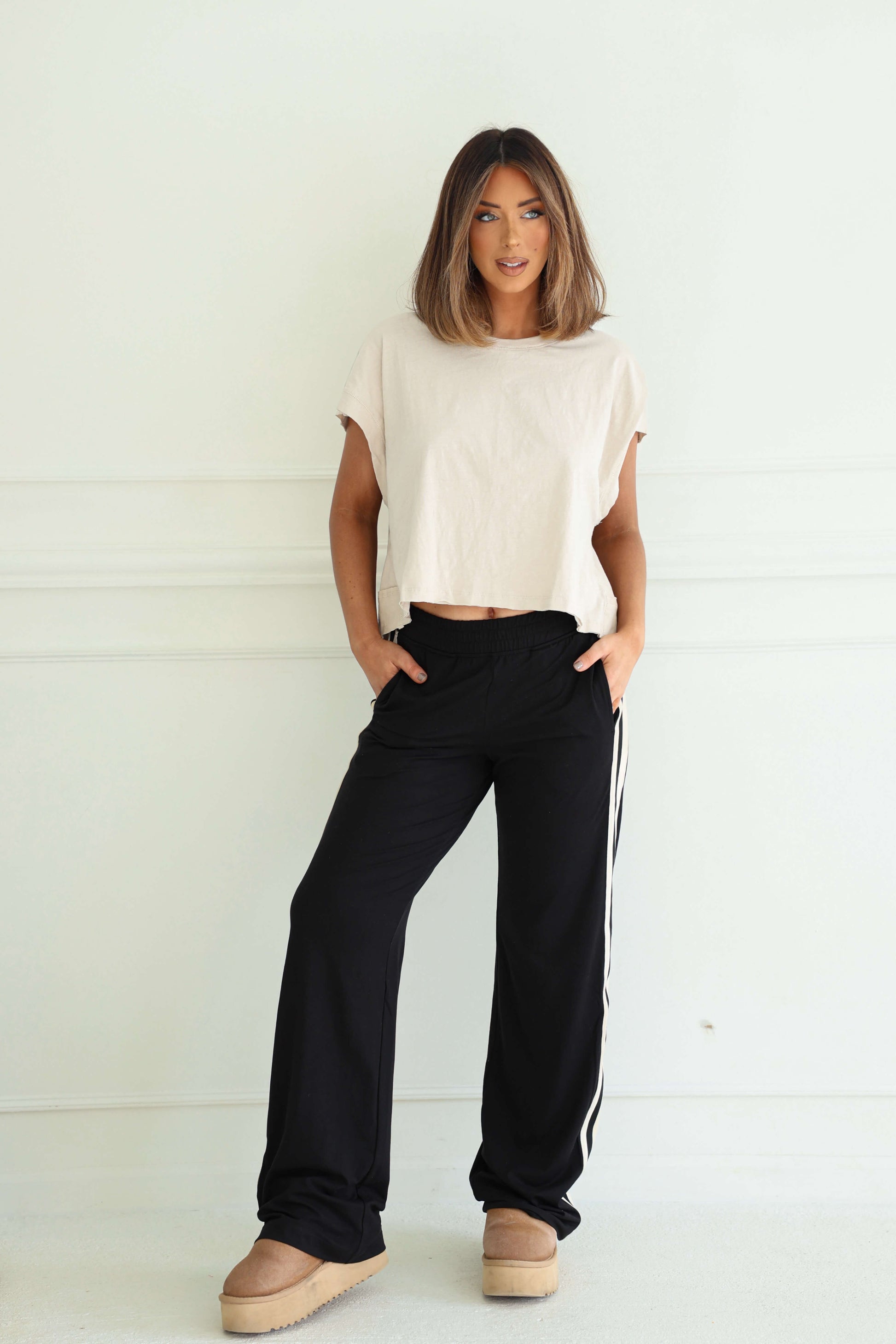 Woman wearing Sporty Black Side Stripe Pants, a beige top, and tan platform shoes while posing against a white wall.