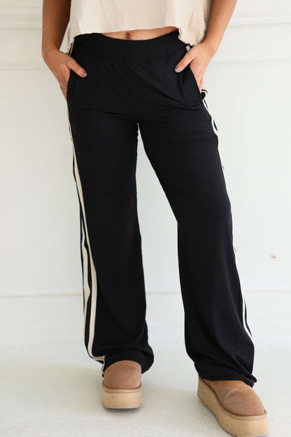 Wearing Sporty Black Side Stripe Pants, a white top, and tan platform slippers, a person stands with hands in pockets.