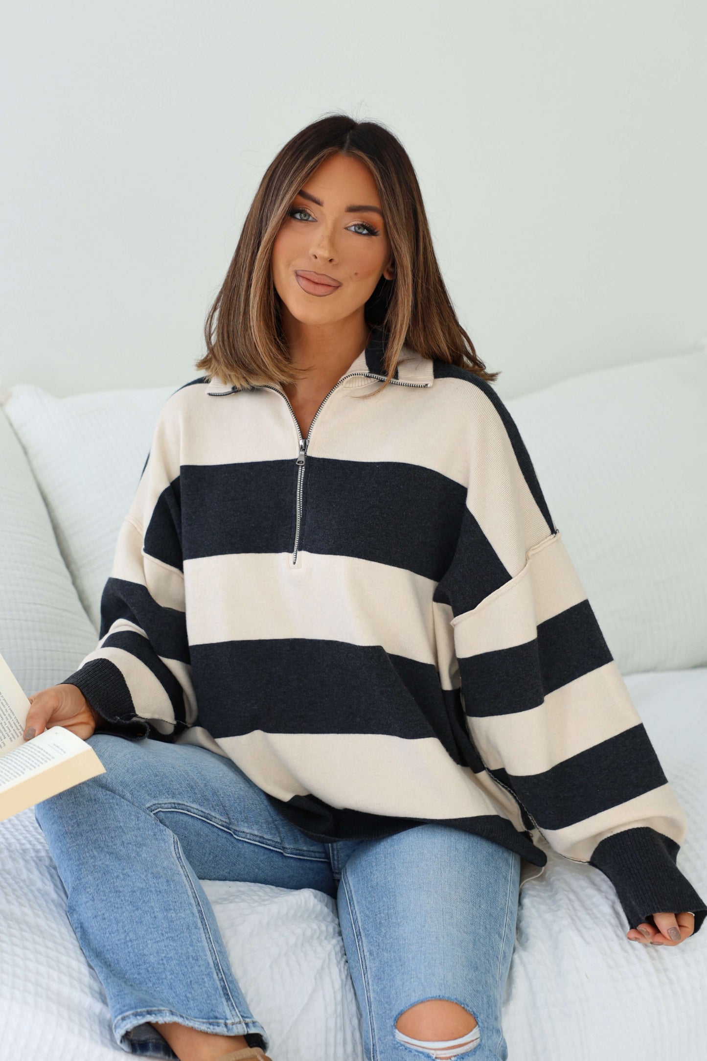 Woman with shoulder-length hair in an Ivory and Charcoal Varsity Stripe Half Zip Rugby Pullover sits on a bed, reading and smiling.