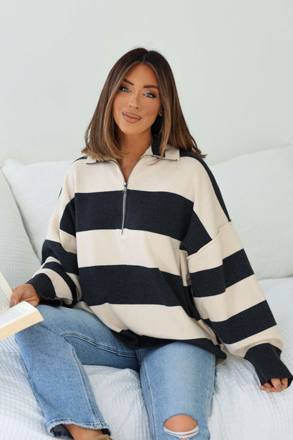 Woman with shoulder-length hair in an Ivory and Charcoal Varsity Stripe Half Zip Rugby Pullover sits on a bed, reading and smiling.