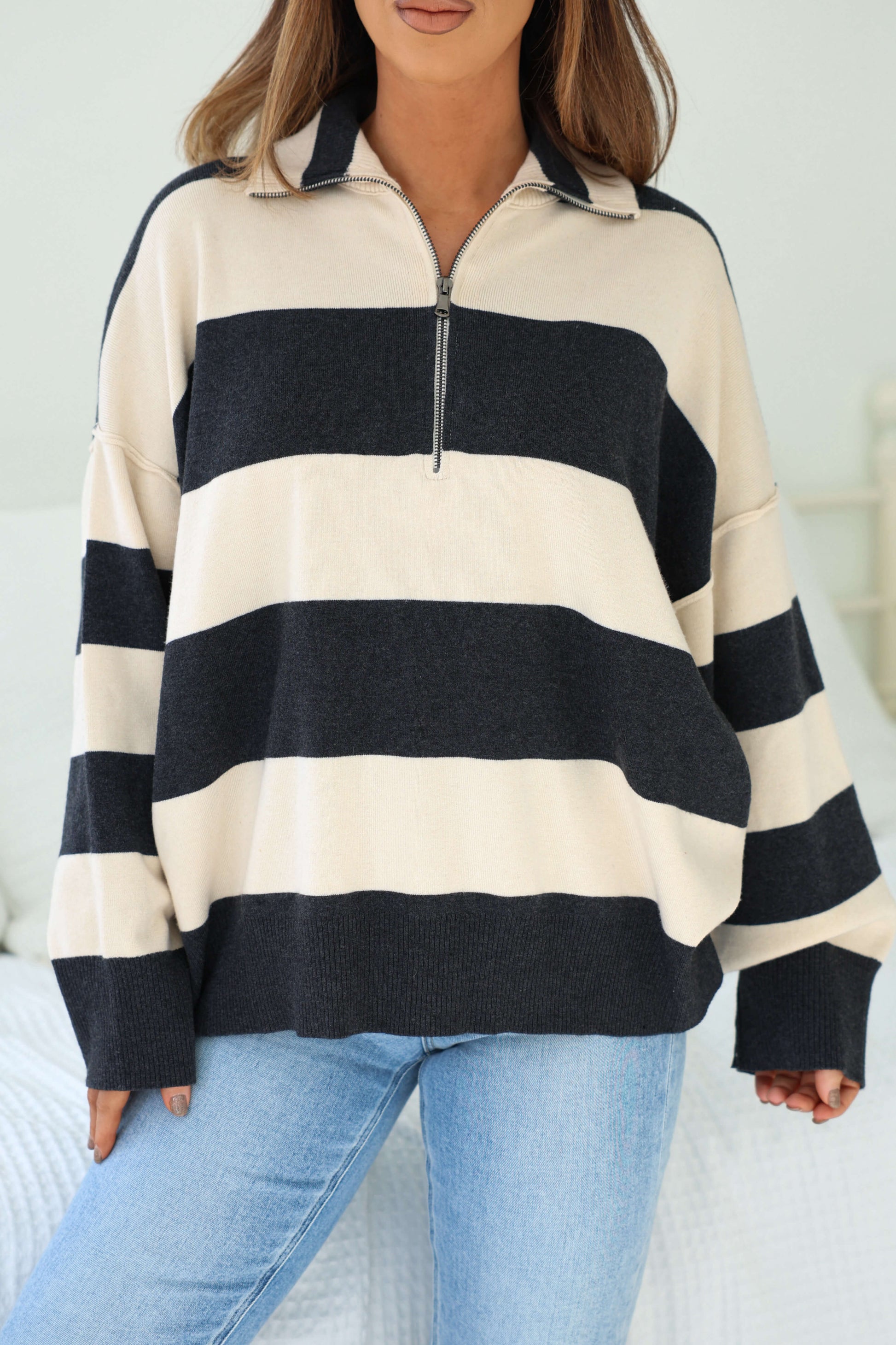 A woman indoors wears an Ivory and Charcoal Varsity Stripe Half Zip Rugby Pullover with light blue jeans.
