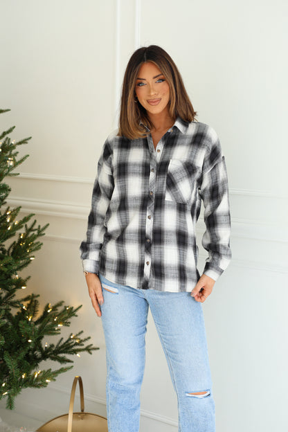 A woman in the Madelyn Charcoal Plaid Flannel Shirt and light blue jeans stands indoors beside a decorated Christmas tree.