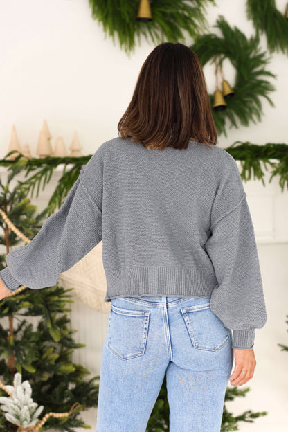A woman in a Grey Waffle Knit Open Front Cardigan and light blue jeans stands with her back to the camera, among festive greenery.