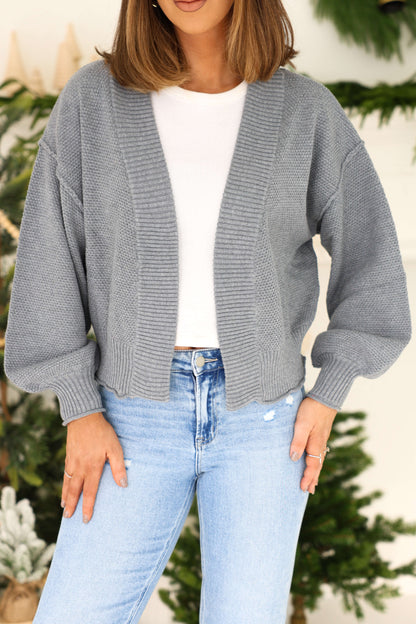 A woman in a Grey Waffle Knit Open Front Cardigan over a white shirt and light blue jeans, standing by a festive green background.