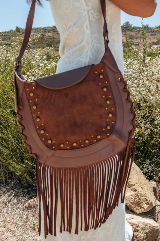 A woman in a white dress holds the Outlaw Fringe Shoulder Bag - Brown, standing outdoors among greenery.