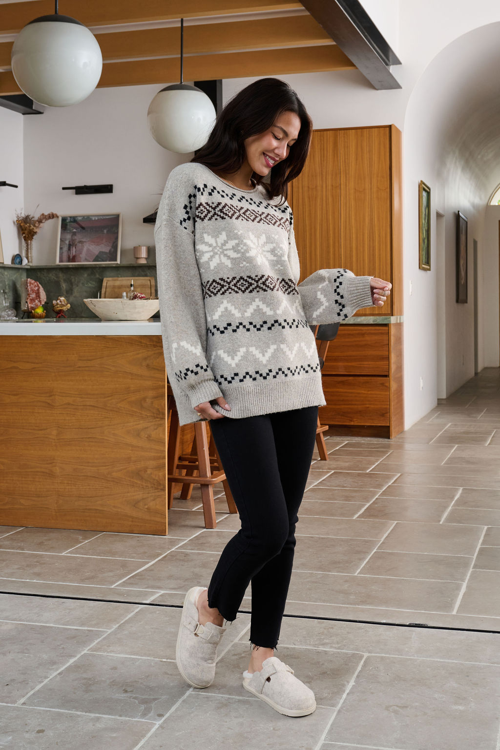 A woman in an oversized Stone Grey Snowflake Sweater smiles in a modern kitchen.