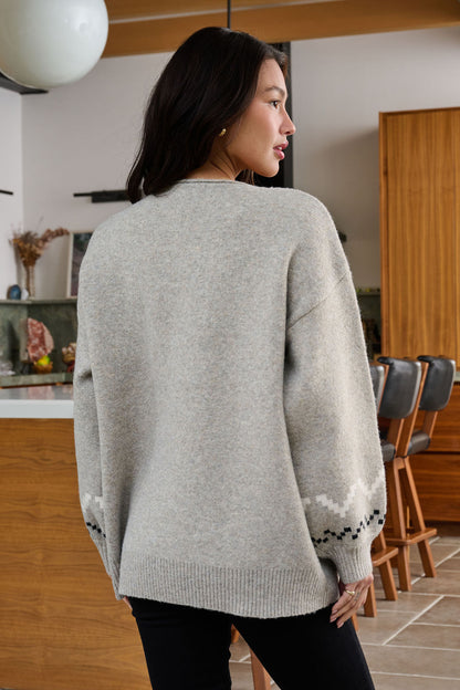 A woman with long dark hair wears a Stone Grey Snowflake Sweater in a modern kitchen, its winter pattern visible from behind.