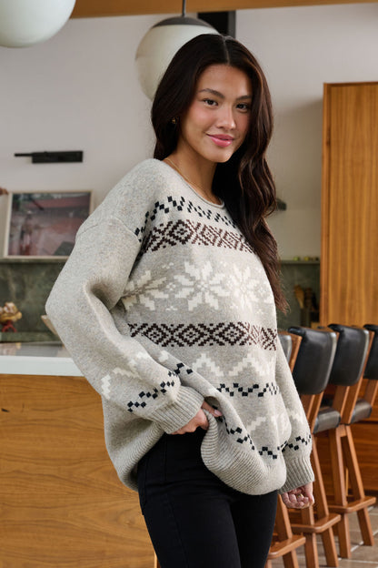A woman smiles indoors with one hand in her pocket, wearing the oversized Stone Grey Snowflake Sweater and black pants.