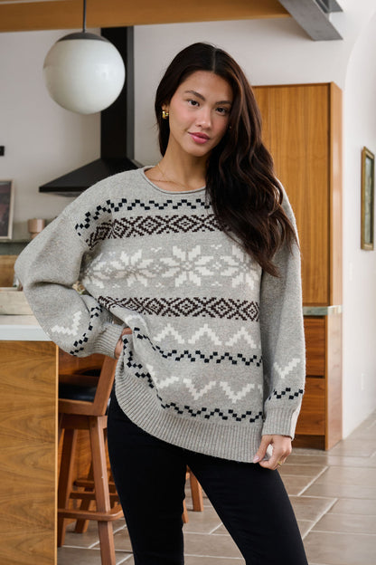 A woman poses casually indoors in a Stone Grey Snowflake Sweater and black pants in a modern kitchen.