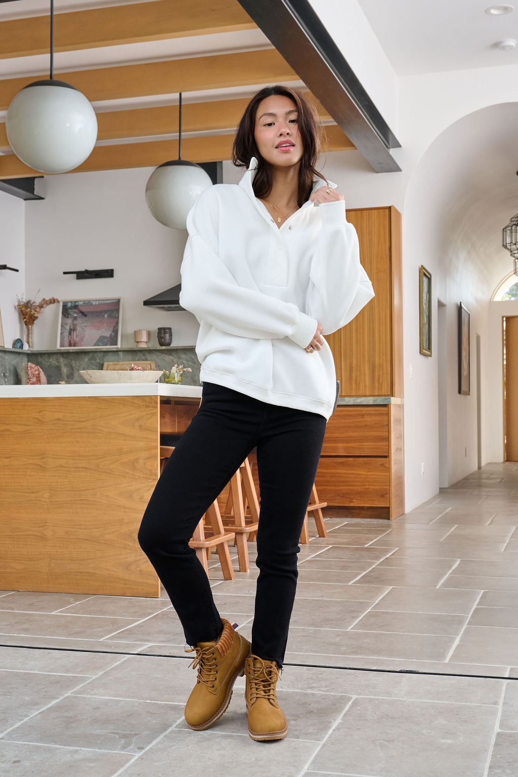 A woman in a Magnolia exclusive White Collared Button Up Pullover Sweatshirt, black pants, and tan boots poses in a modern kitchen.