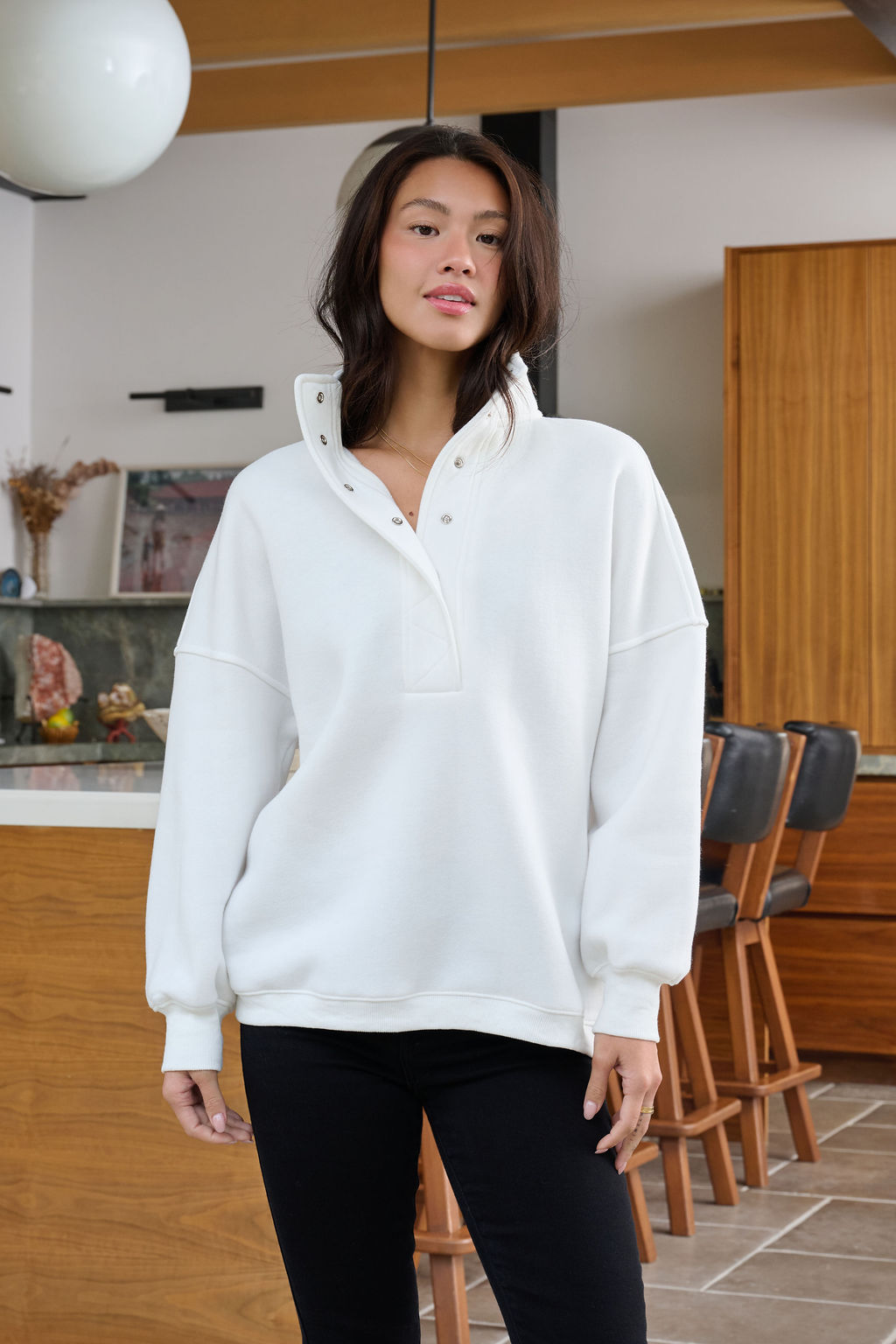 A woman stands indoors in a White Collared Button Up Pullover Sweatshirt and black pants, with wooden furniture and bar stools behind her.