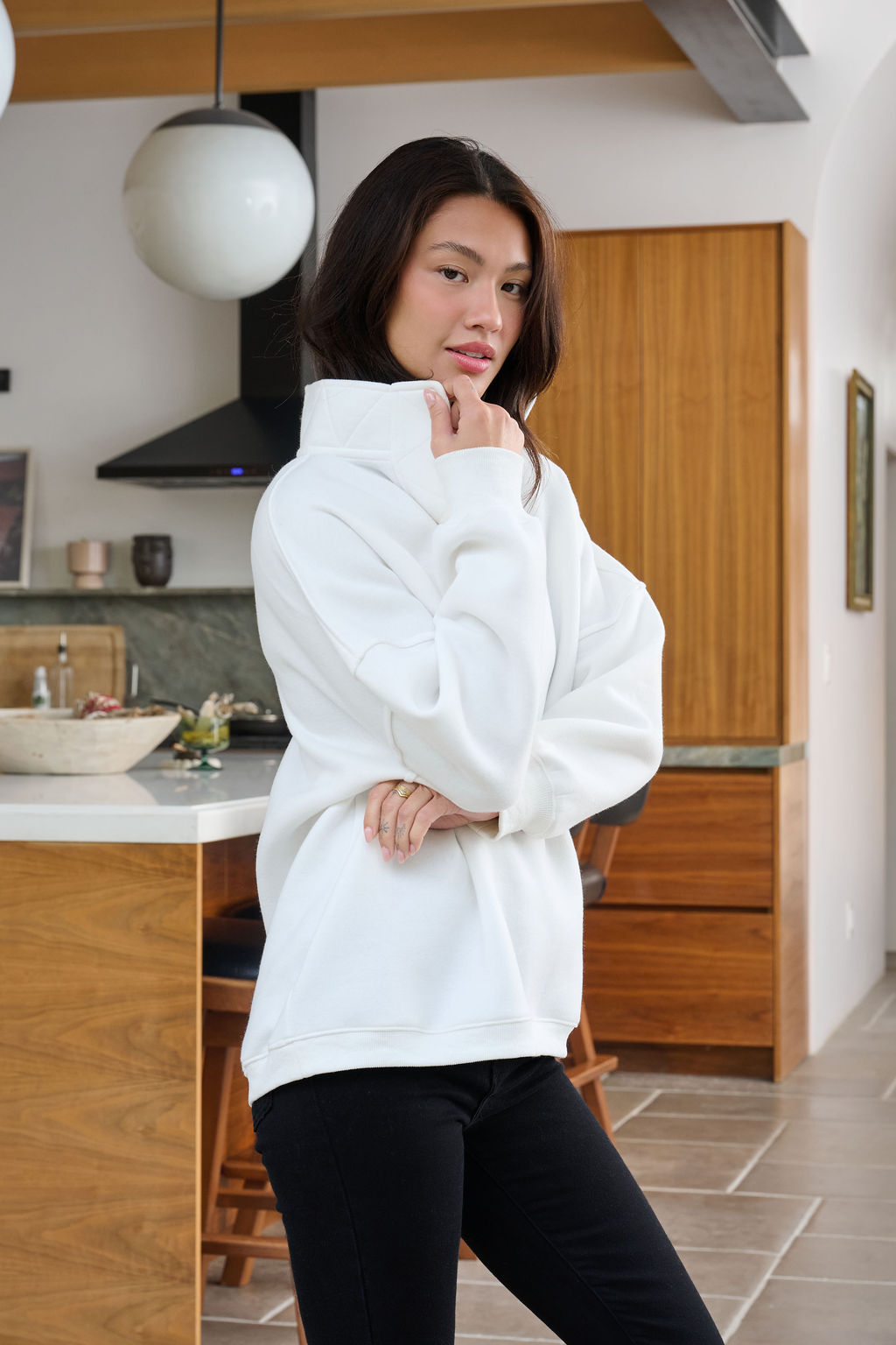 A woman models the White Collared Button Up Pullover Sweatshirt in a modern kitchen, gently touching her collar and smiling.