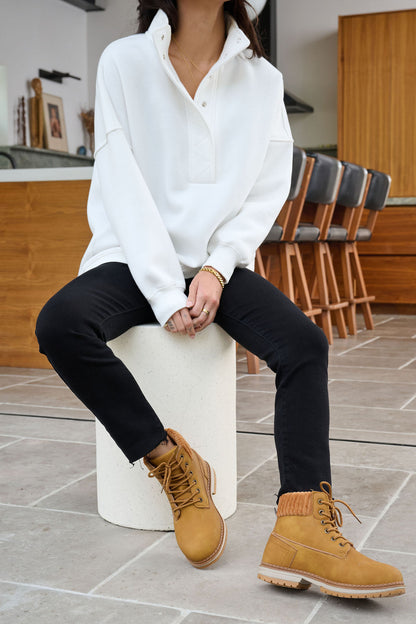 A woman in a White Collared Button Up Pullover Sweatshirt, black pants, and tan boots sits on a stool in a modern kitchen.
