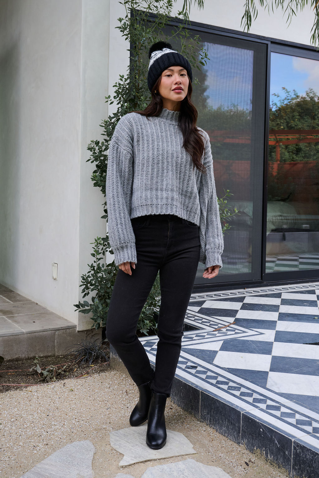 A woman in a Grey Wide Sleeve Ribbed Mock Neck Sweater, black pants, and a beanie stands outside on a tiled patio by glass doors.